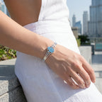 Woman wearing powder blue Hamsa bangle on wrist, Dubai city towers visible in background, silver cuff detail