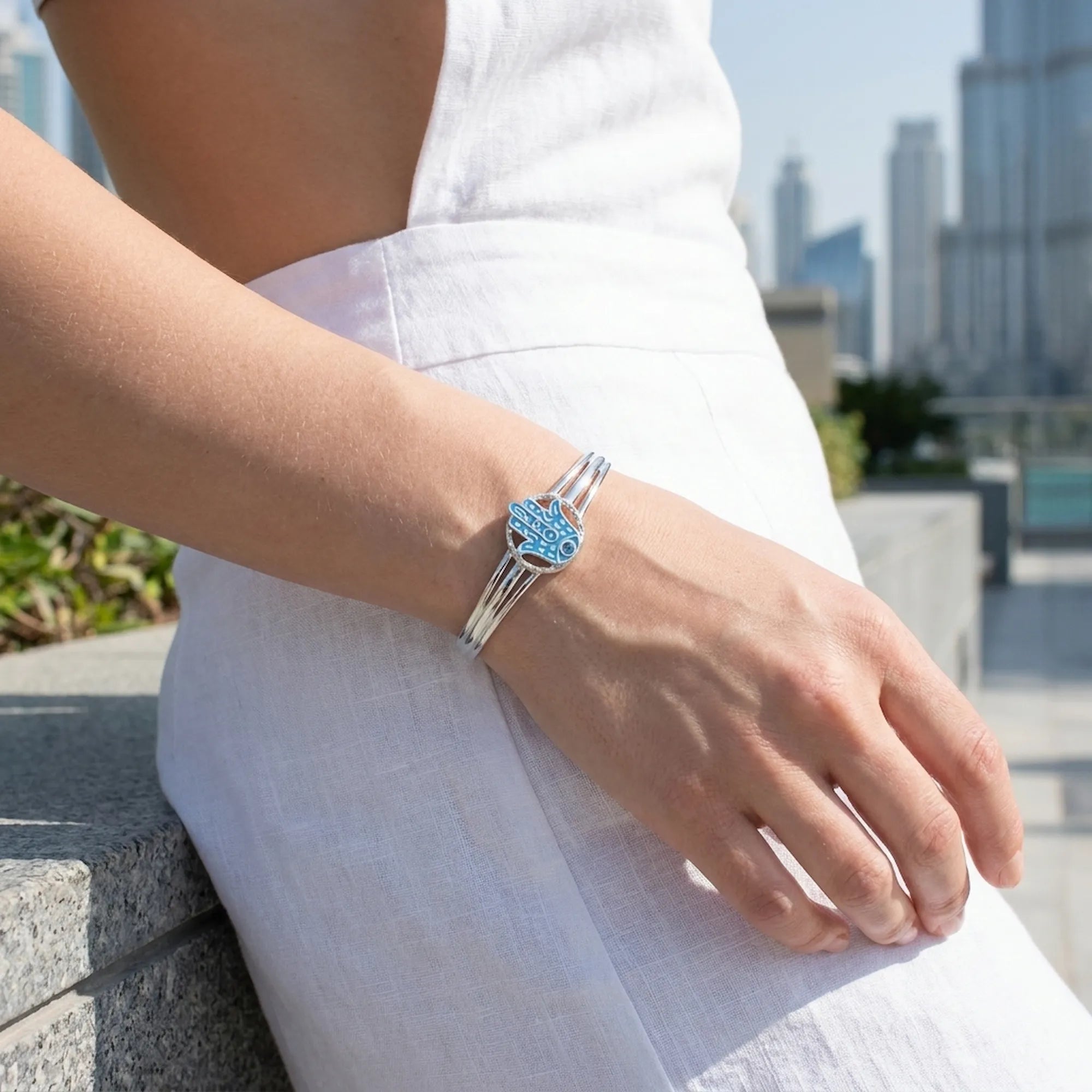 Woman wearing powder blue Hamsa bangle on wrist, Dubai city towers visible in background, silver cuff detail
