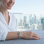 Woman wearing teal Hamsa bangle on wrist, Dubai city skyline visible through glass in background