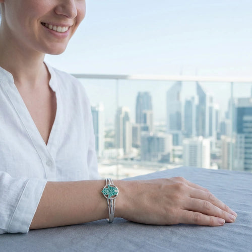 Woman wearing teal Hamsa bangle on wrist, Dubai city skyline visible through glass in background