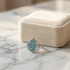 Silver hamsa ring with powder blue enamel and a blue topaz eye, resting on marble beside a cream jewelry box.
