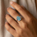 Silver hamsa ring with powder blue enamel and blue topaz worn on the finger, hand against soft fabric.
