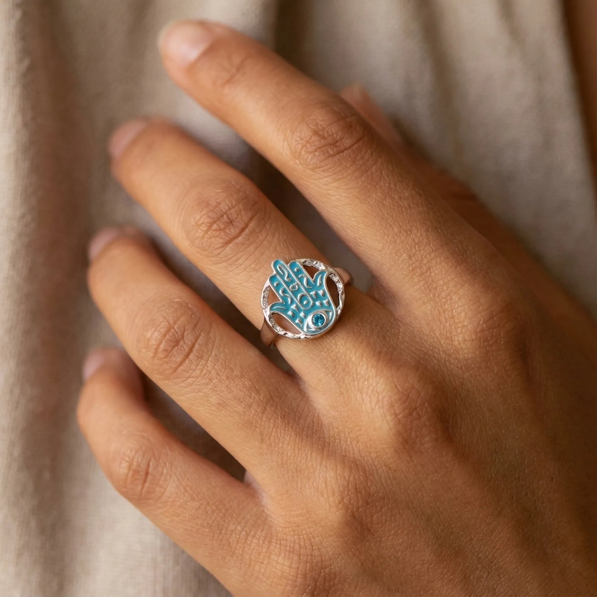 Silver hamsa ring with powder blue enamel and blue topaz worn on the finger, hand against soft fabric.