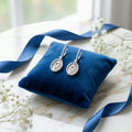 Silver Hamsa drop earrings on blue velvet cushion with white flowers and ribbon — wedding and occasion styling UAE.