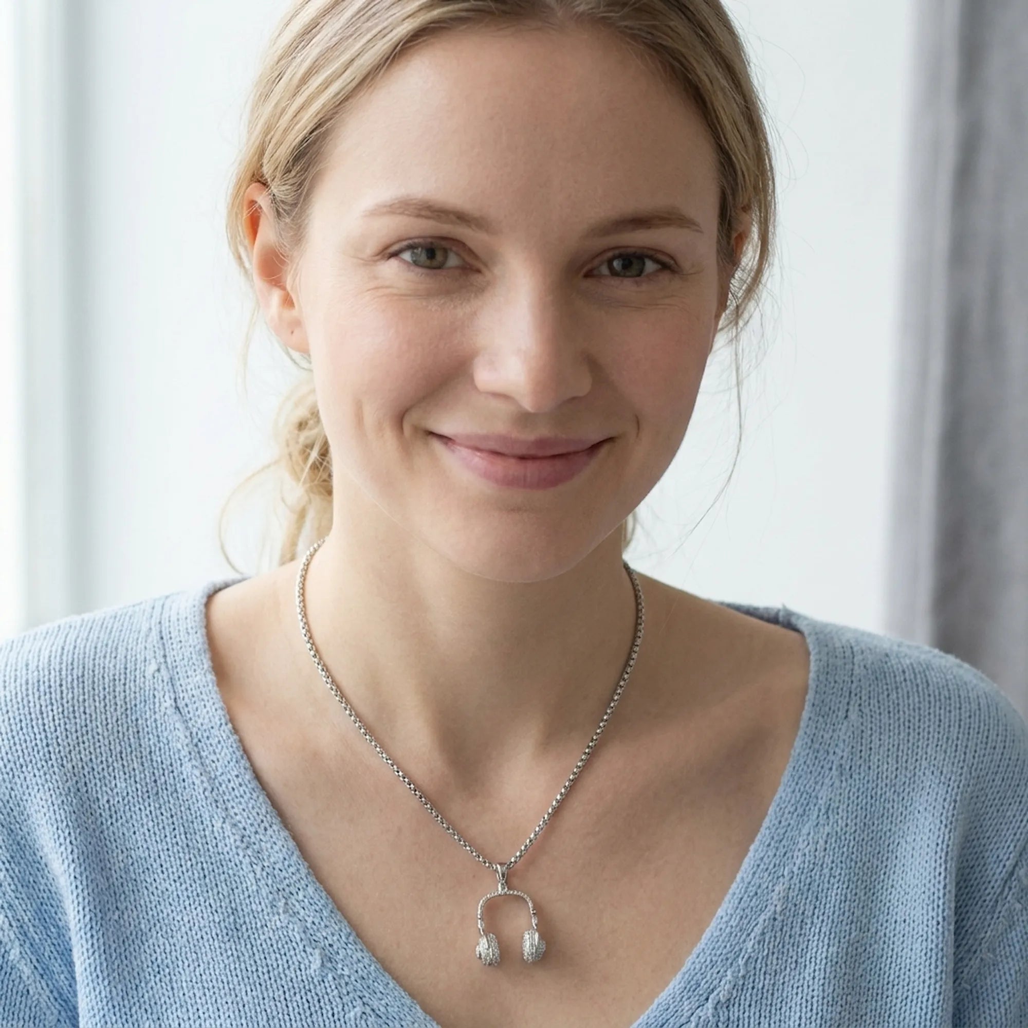 Woman wearing silver DJ headphone pendant necklace on thick box chain with V-neck blue sweater, neutral background