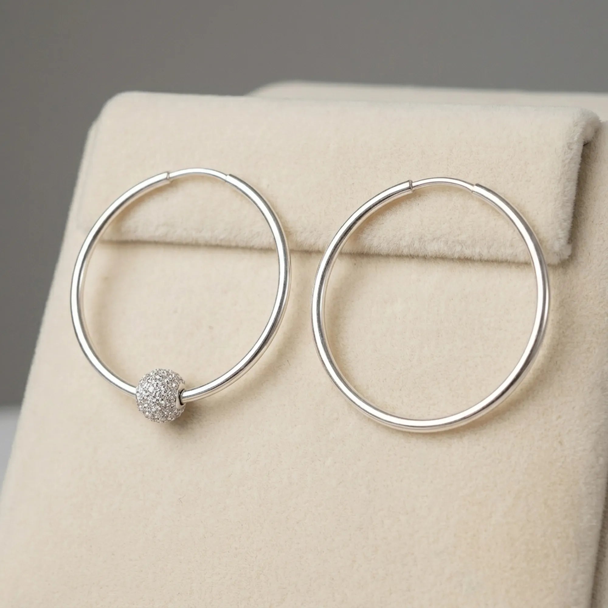 Silver hoop earrings on cream velvet display stand one showing CZ pave bead position and one showing clean hoop without bead