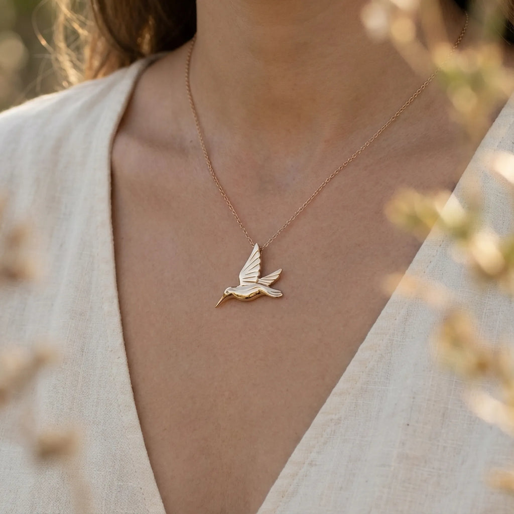 Gold hummingbird pendant necklace worn over a cream wrap top, blurred garden behind.
