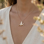 Gold hummingbird pendant necklace worn over a cream wrap top, blurred garden behind.