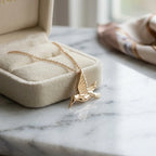 Gold-toned hummingbird pendant necklace resting on marble beside a cream jewelry box and blurred scarf Luna Dura studio.