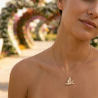 Gold hummingbird pendant on a fine chain worn on the neck, flower arches of Dubai Miracle Garden behind.