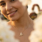 Gold hummingbird pendant necklace worn on the neck close up in soft sunlight, blurred flowers behind.