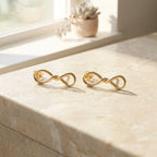 Both gold infinity drop earrings laid flat on marble windowsill with succulent plant in background