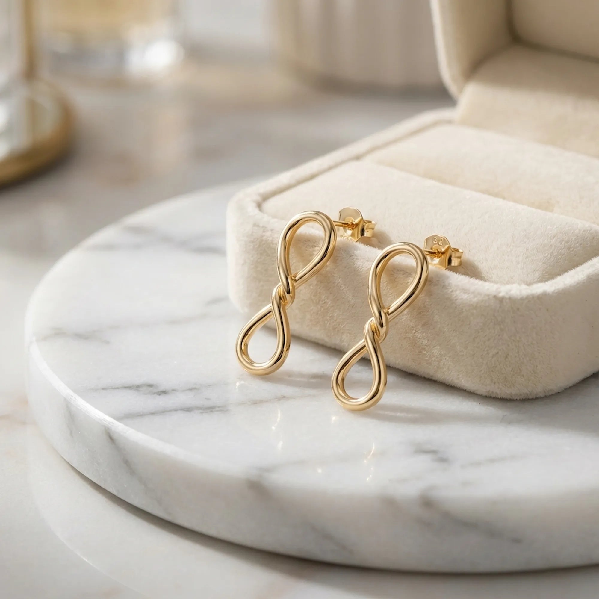 Gold infinity drop earrings resting against cream velvet jewellery box on marble tray
