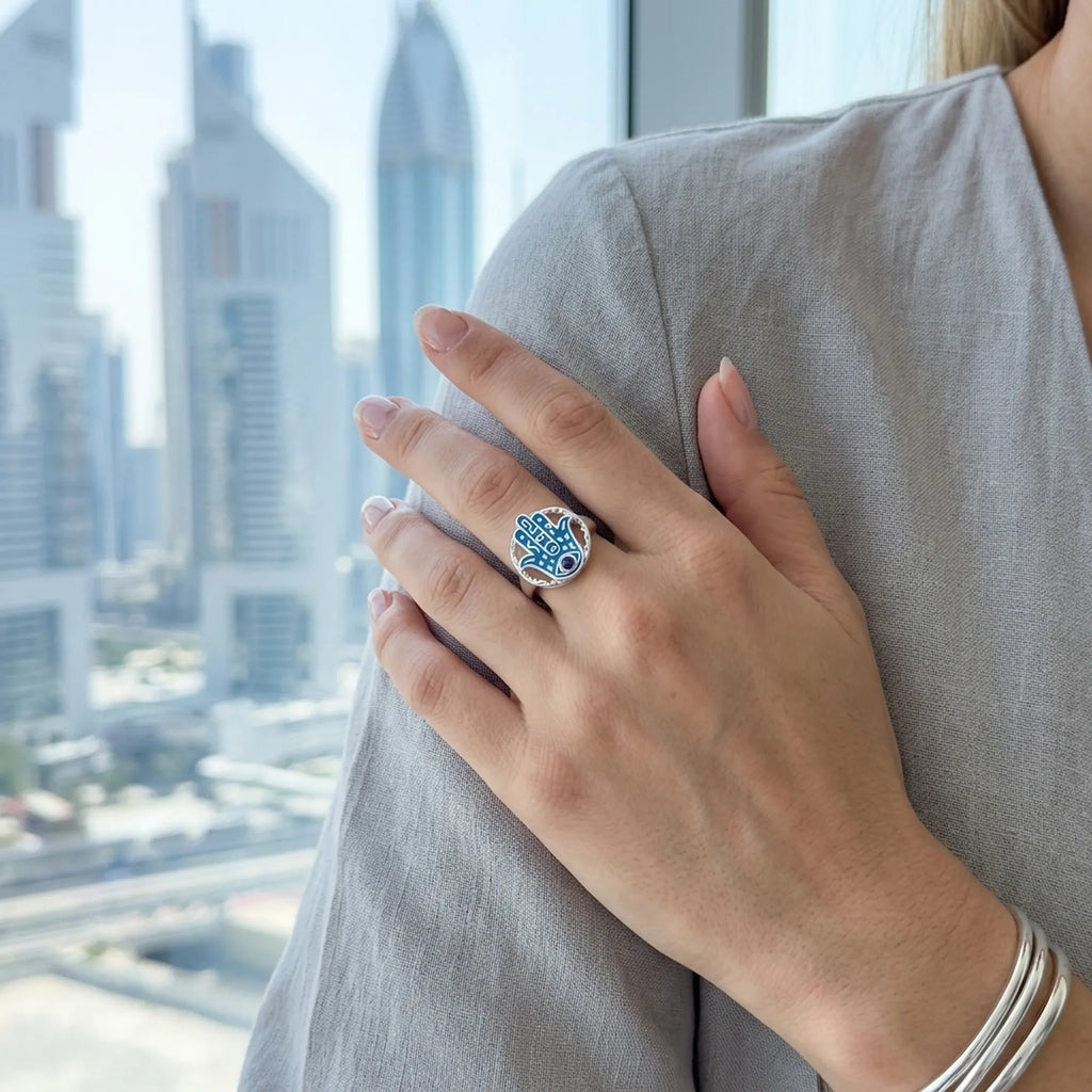 Azure enamel hamsa ring with tanzanite worn on the hand, Jumeirah Emirates Towers visible through the window behind.