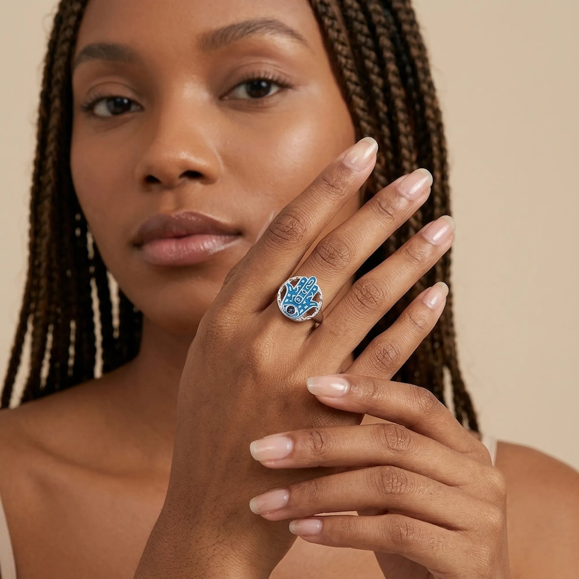 Azure enamel hamsa ring with a tanzanite eye worn on the finger, model with olive skin color against a soft beige background.