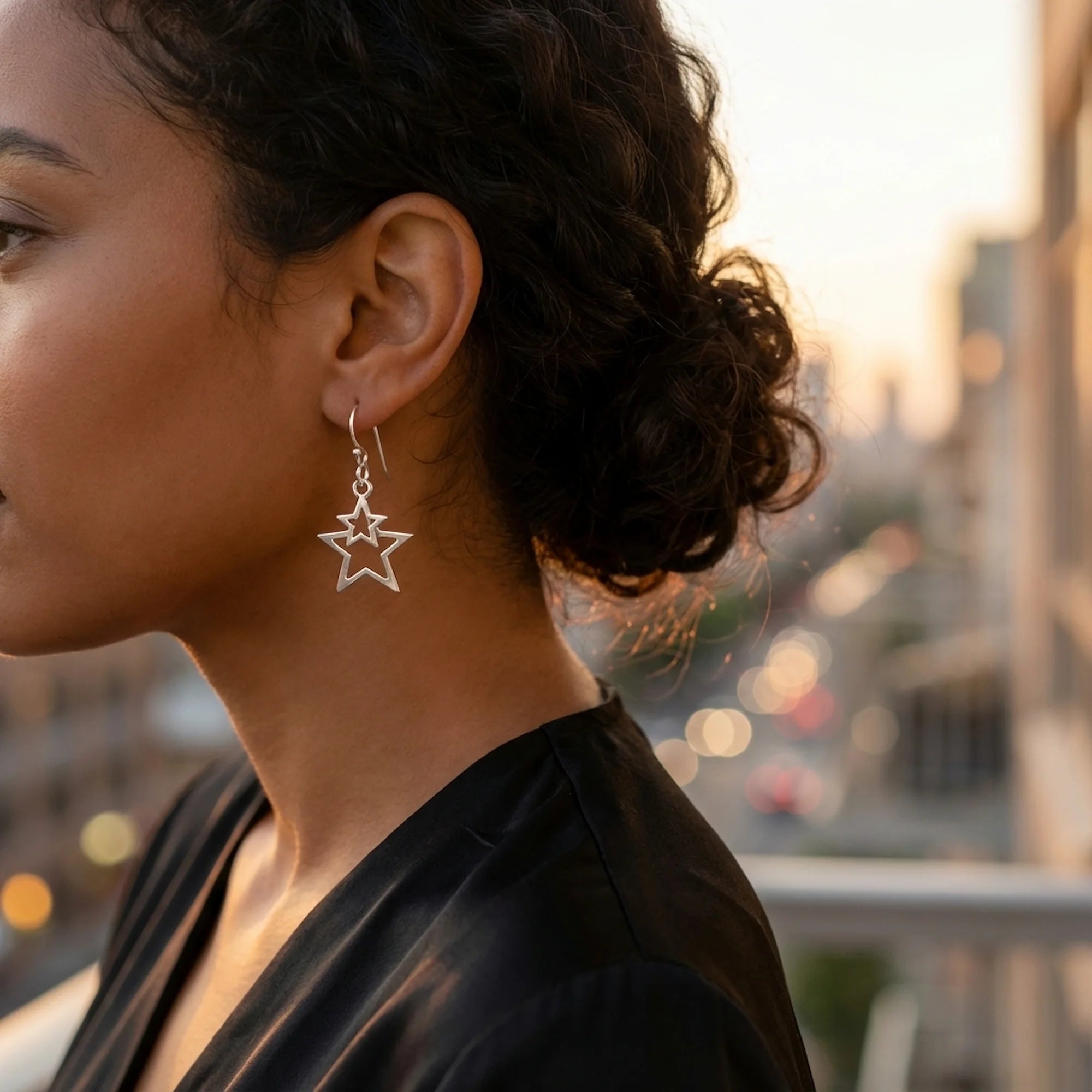 Woman wearing pure-silver plated double star drop earrings outdoors in Dubai with a blurred city sunset background.