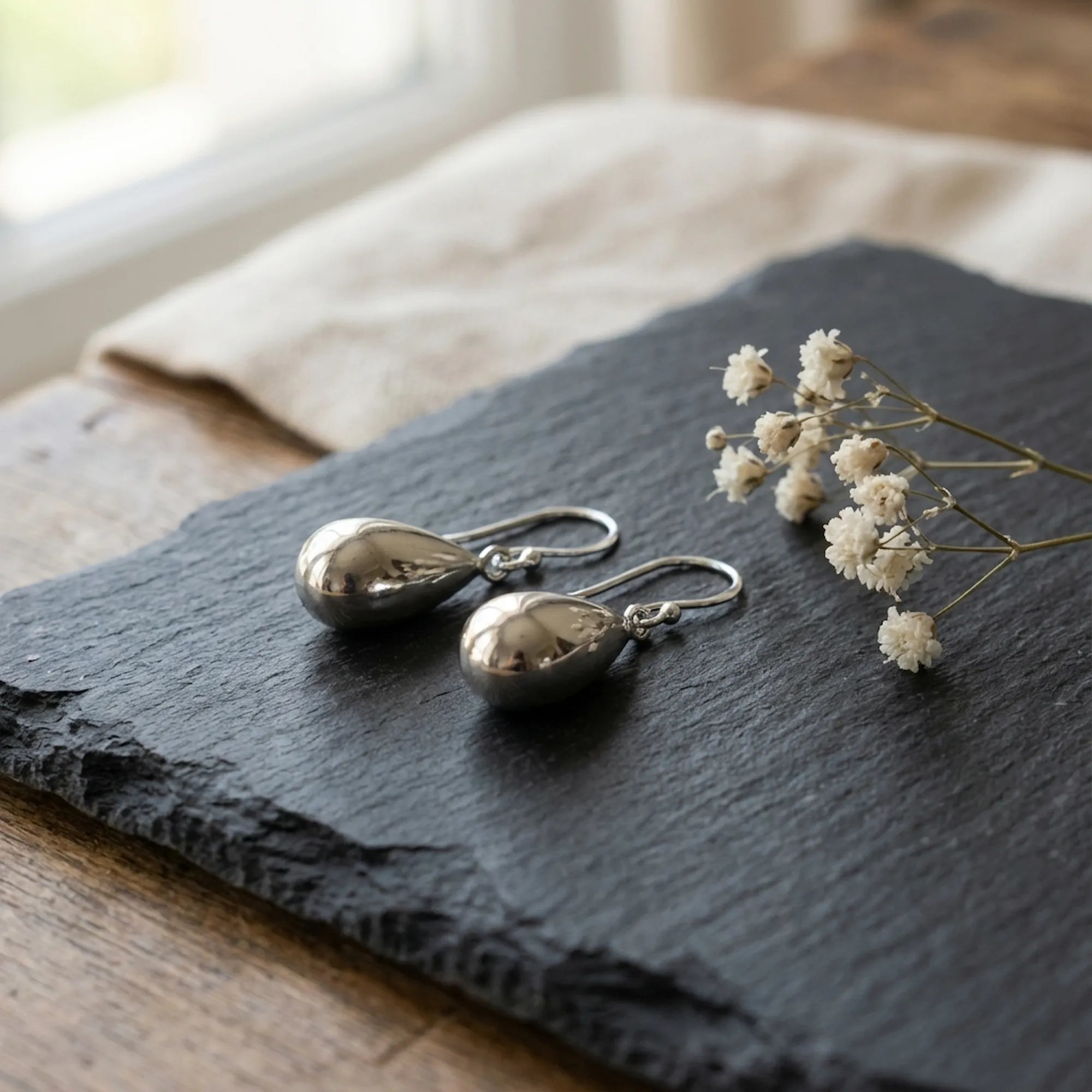 Molten Drop silver teardrop earrings on dark slate with dried white flowers — minimalist flat lay 925 sterling silver Dubai