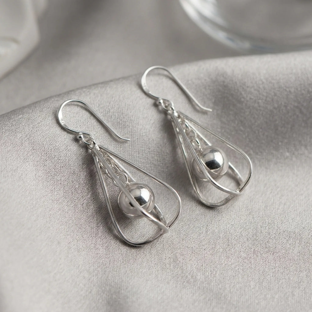 Pair of polished silver orb drop earrings laid on grey fabric showing frame structure and suspended sphere detail