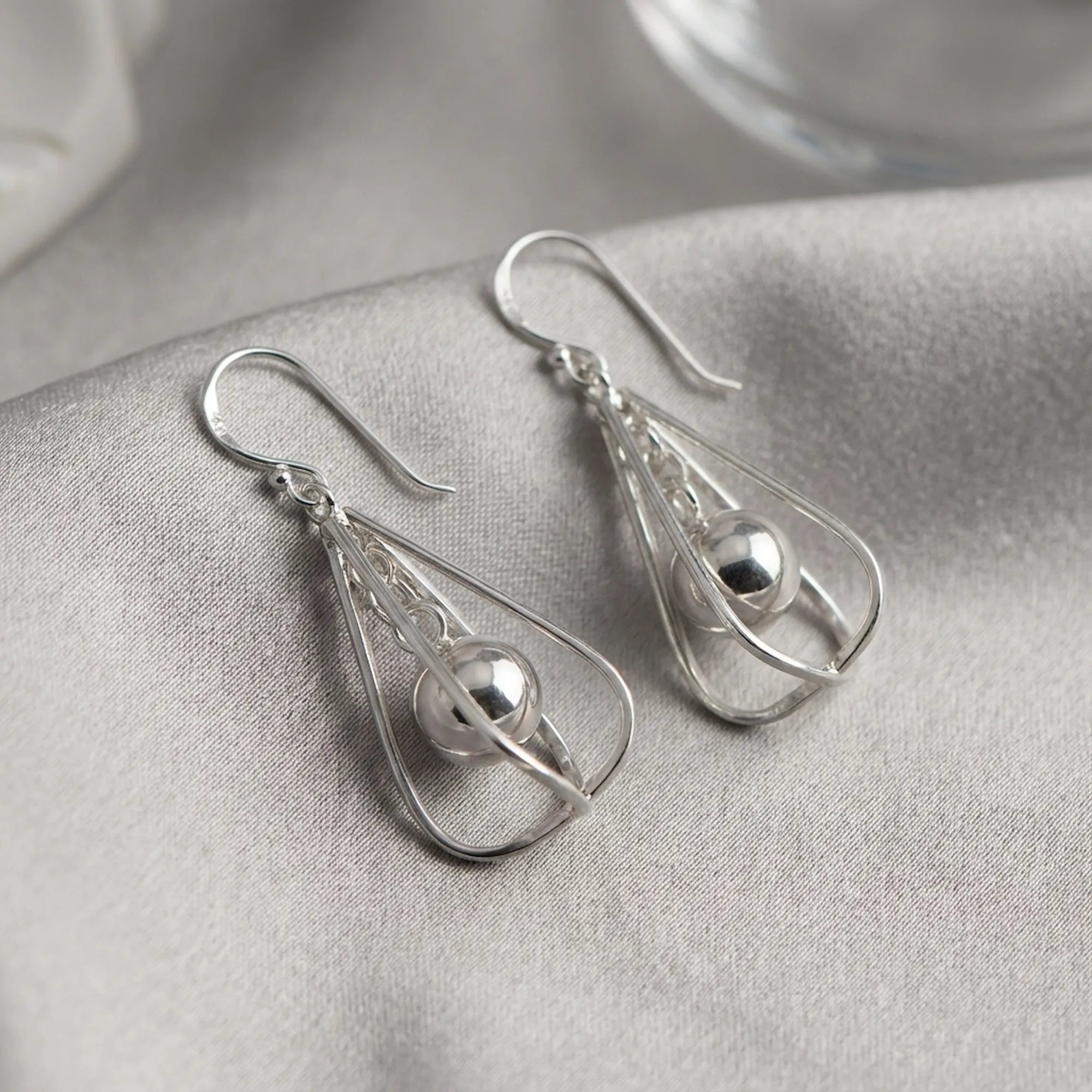 Pair of polished silver orb drop earrings laid on grey fabric showing frame structure and suspended sphere detail