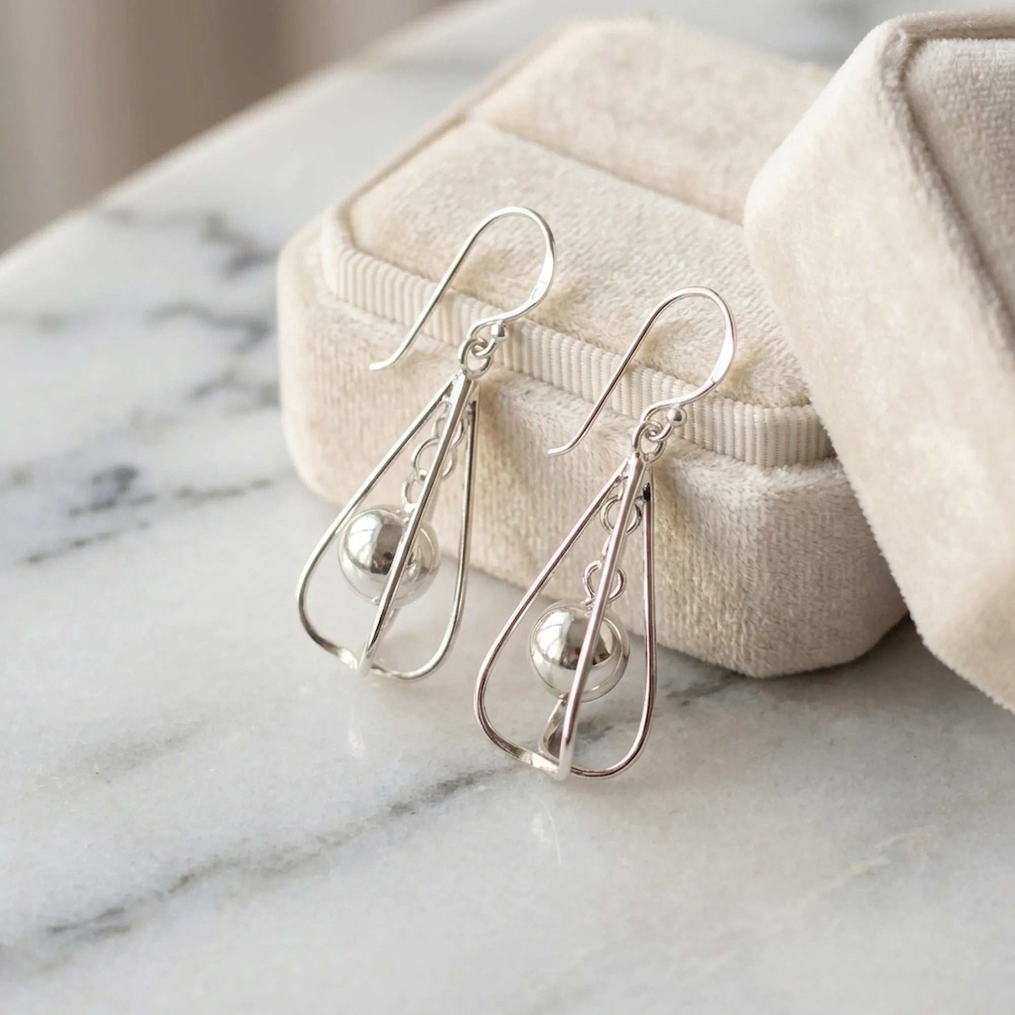 Silver orb drop earrings leaning against open Luna Dura cream velvet gift box on white marble surface