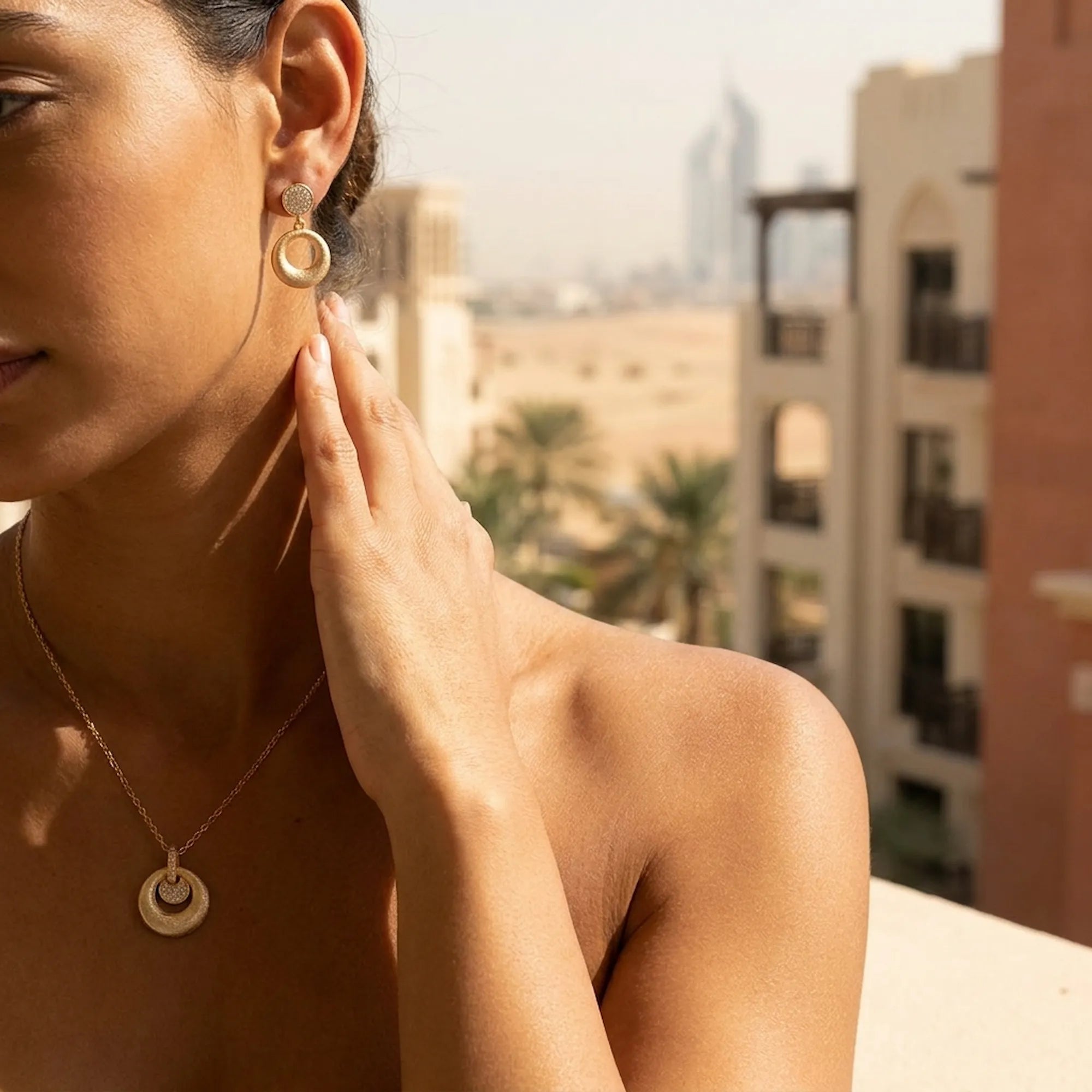 18K gold circle drop earrings and orbiting disc pendant worn in Dubai — city skyline and palm trees background