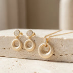 Brushed gold open ring drop earrings and orbiting disc pendant necklace displayed on travertine stone surface