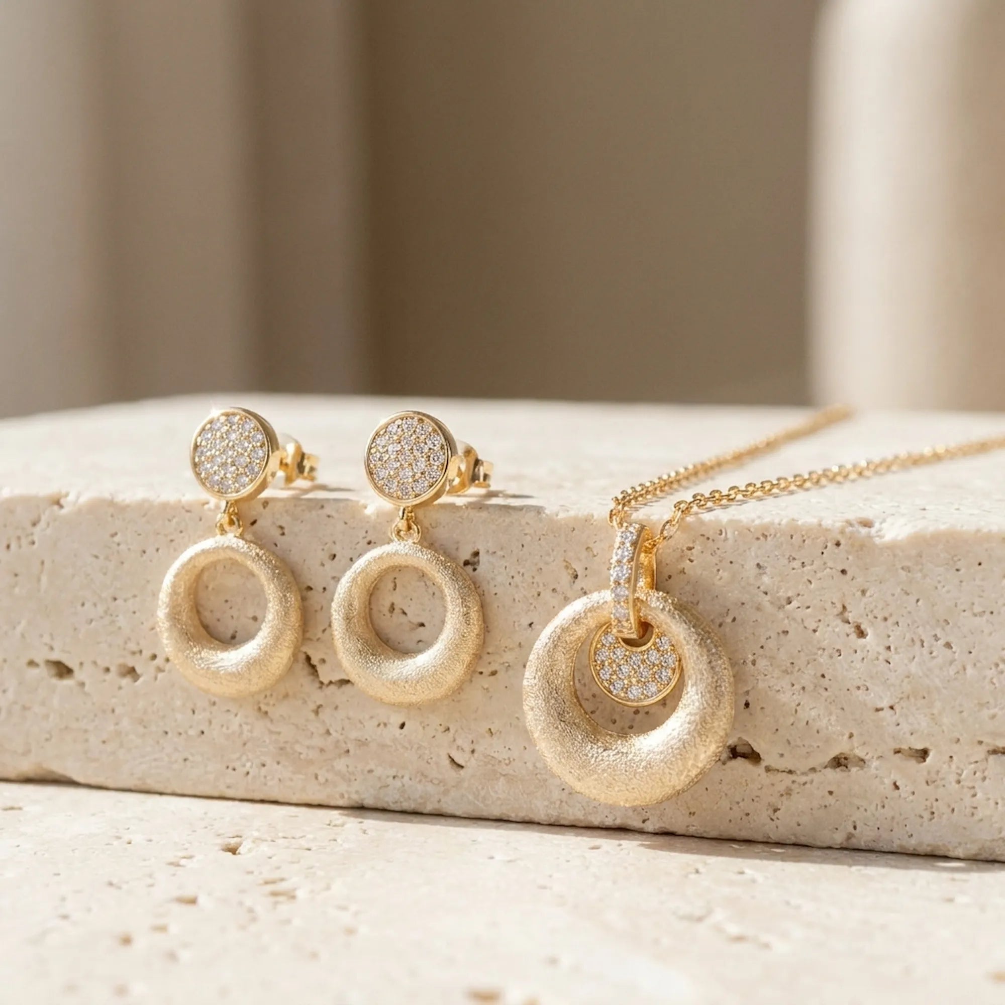 Brushed gold open ring drop earrings and orbiting disc pendant necklace displayed on travertine stone surface