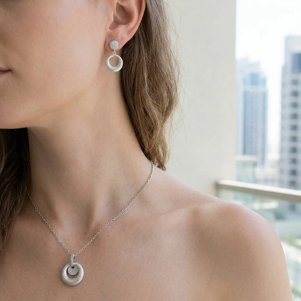 Brushed silver orbiting disc drop earring and pendant at neckline — cool skin model with Dubai skyline background