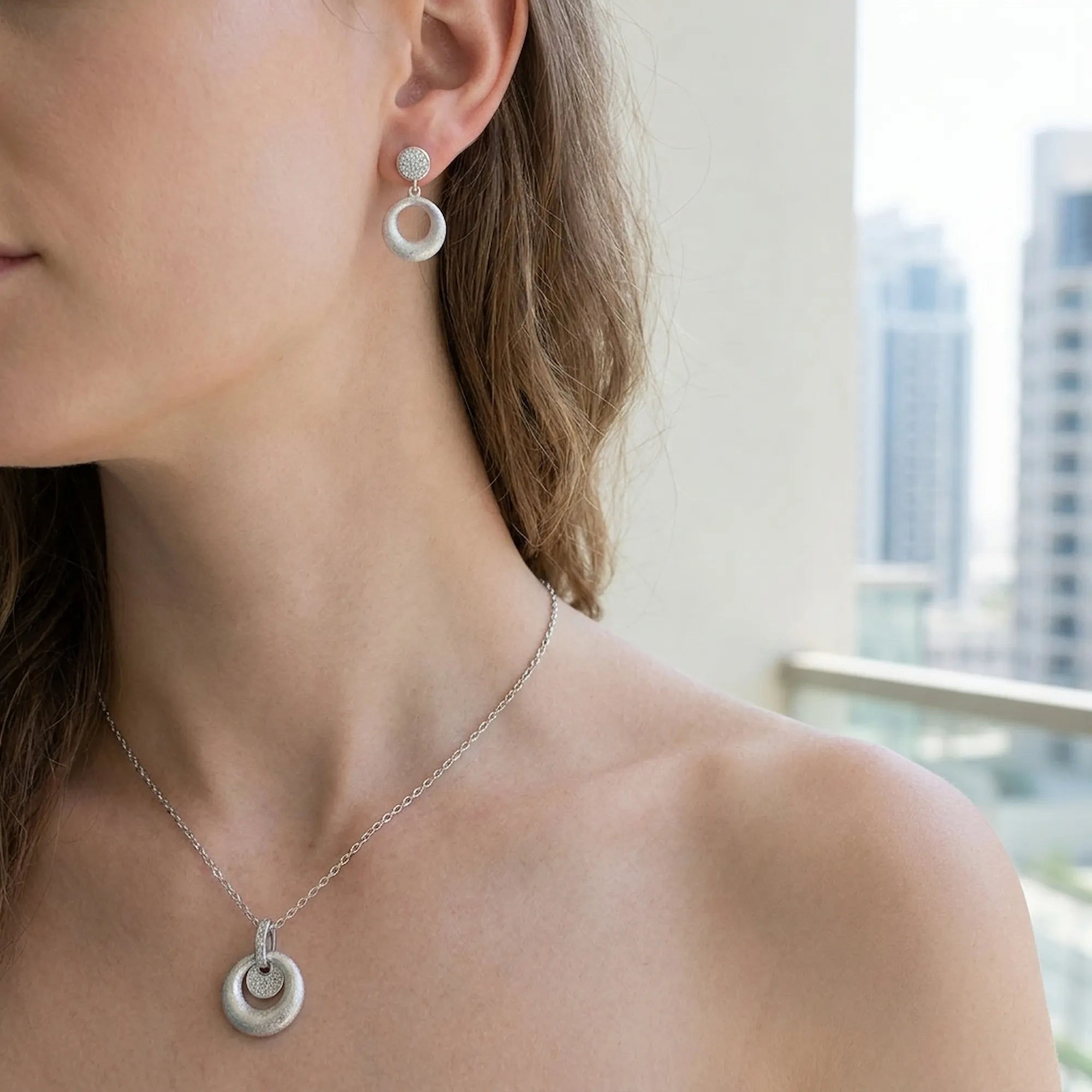 Brushed silver orbiting disc drop earring and pendant at neckline — cool skin model with Dubai skyline background