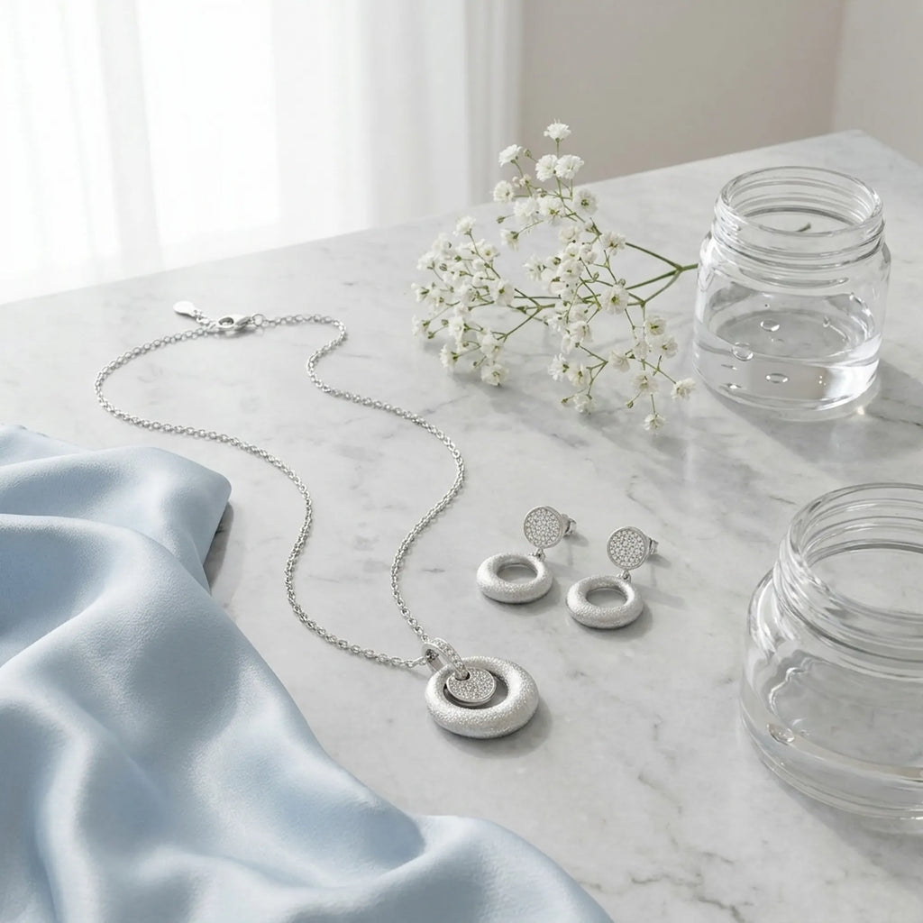 Silver orbiting disc pendant necklace and earring set on marble with blue silk and white flowers — cool editorial styling