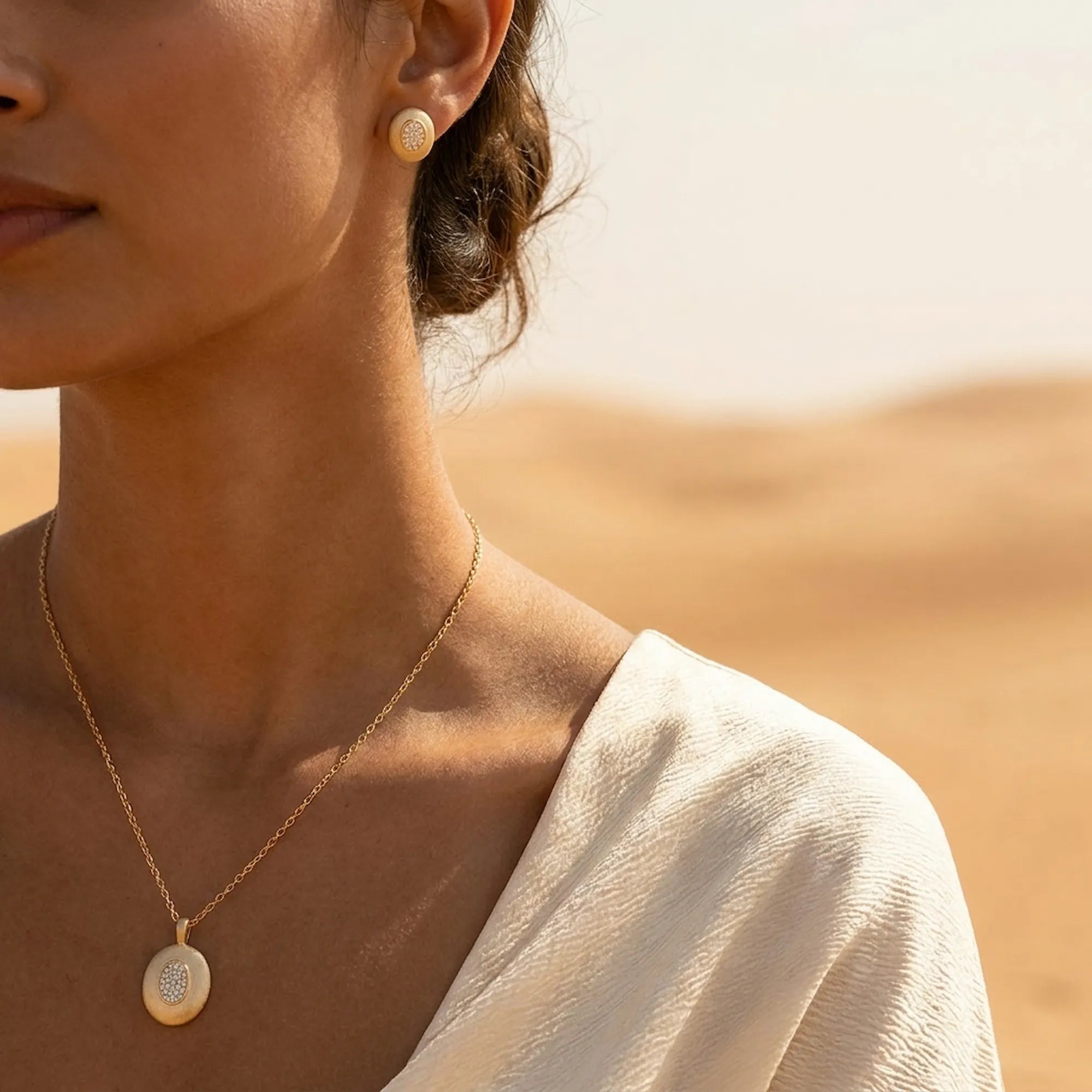 Model wearing brushed gold oval earrings and pendant necklace with sandy desert dunes in background UAE