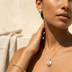 Close-up of brushed gold oval stud earring and oval pendant at neckline on warm olive skin tone