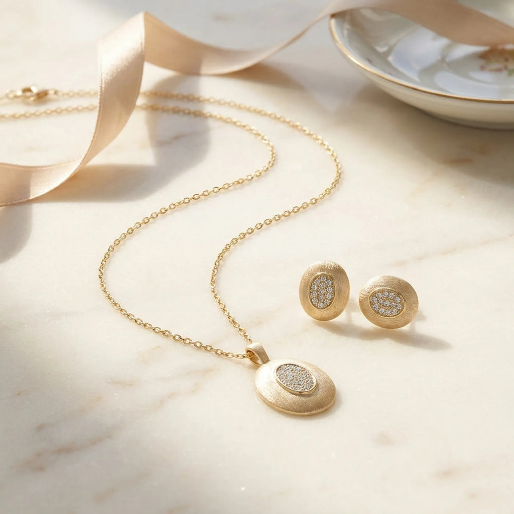 Brushed gold oval pendant and earring set on marble surface with champagne ribbon — gift presentation