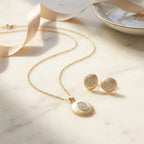 Brushed gold oval pendant and earring set on marble surface with champagne ribbon — gift presentation