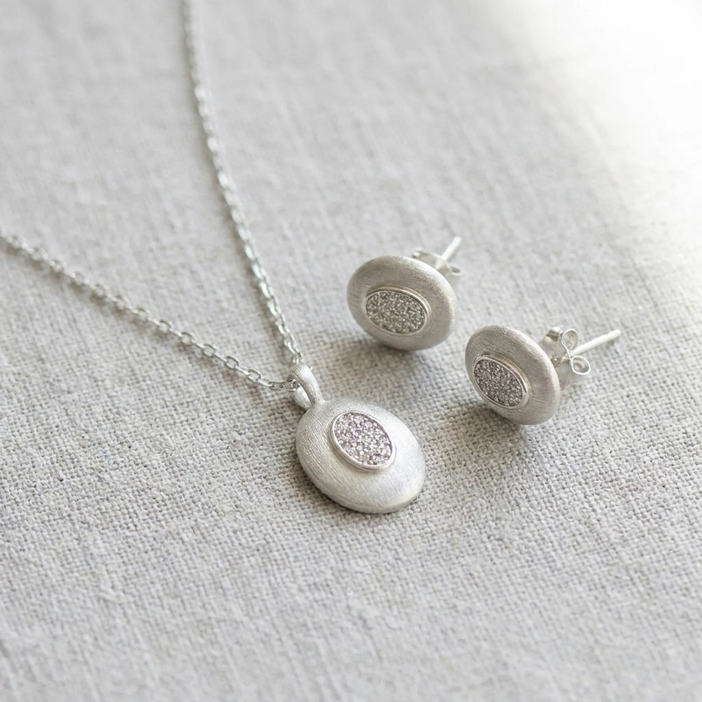 Rhodium silver oval pendant necklace and stud earrings on linen — brushed 925 sterling silver with CZ pavé centre