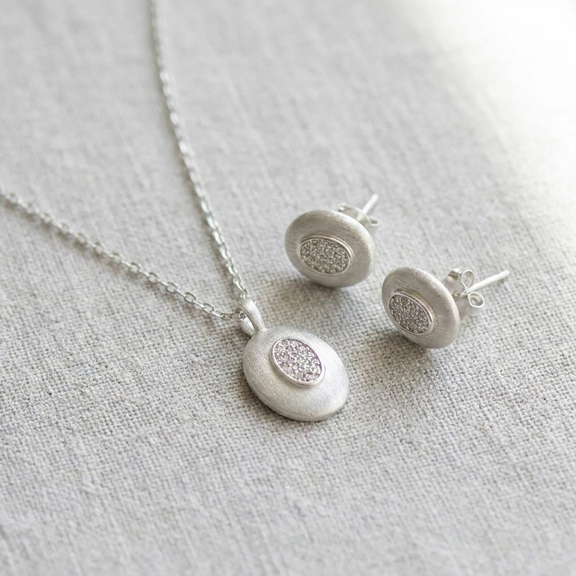 Rhodium silver oval pendant necklace and stud earrings on linen — brushed 925 sterling silver with CZ pavé centre