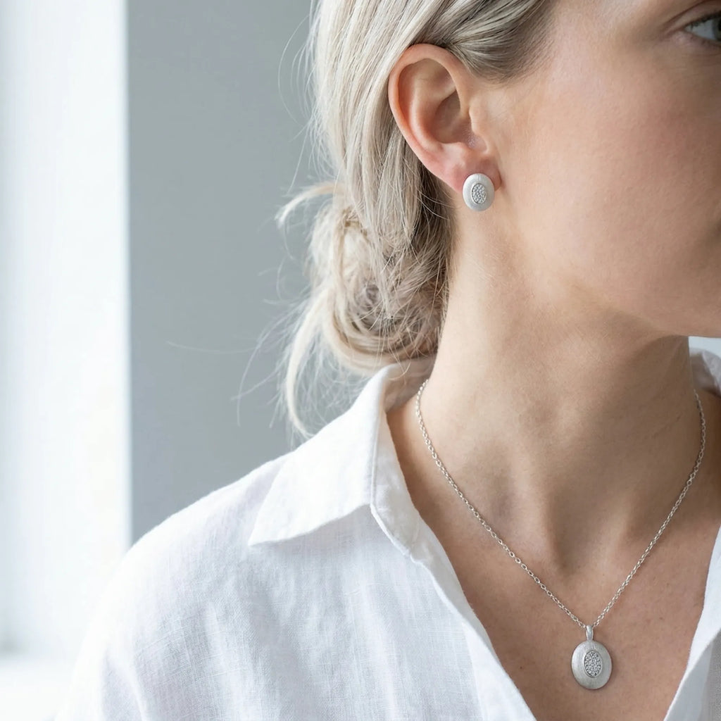 Brushed silver oval pendant necklace and stud earrings on fair skin model with white linen shirt — 925 sterling silver