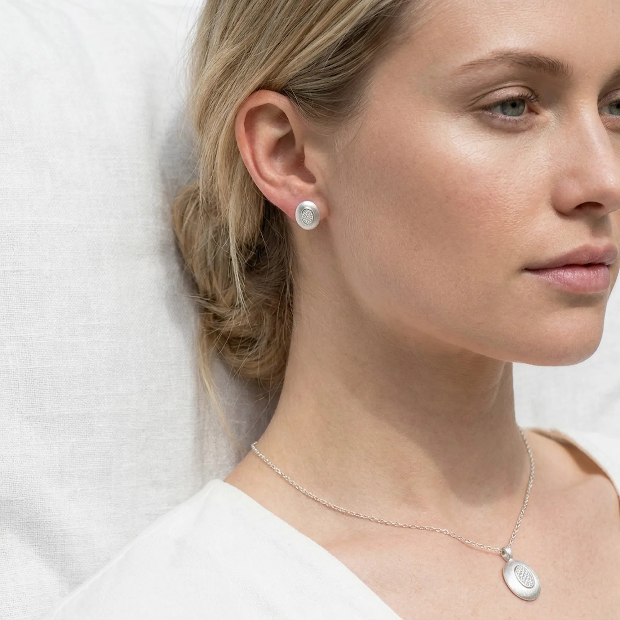 Fair skin model wearing rhodium silver oval earring and pendant necklace against soft white background