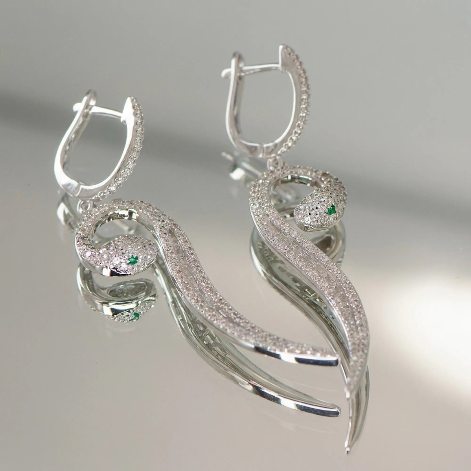 Both paved serpent drop earrings on reflective surface — huggie clicker open, green zircon CZ eyes and baguette spine detail