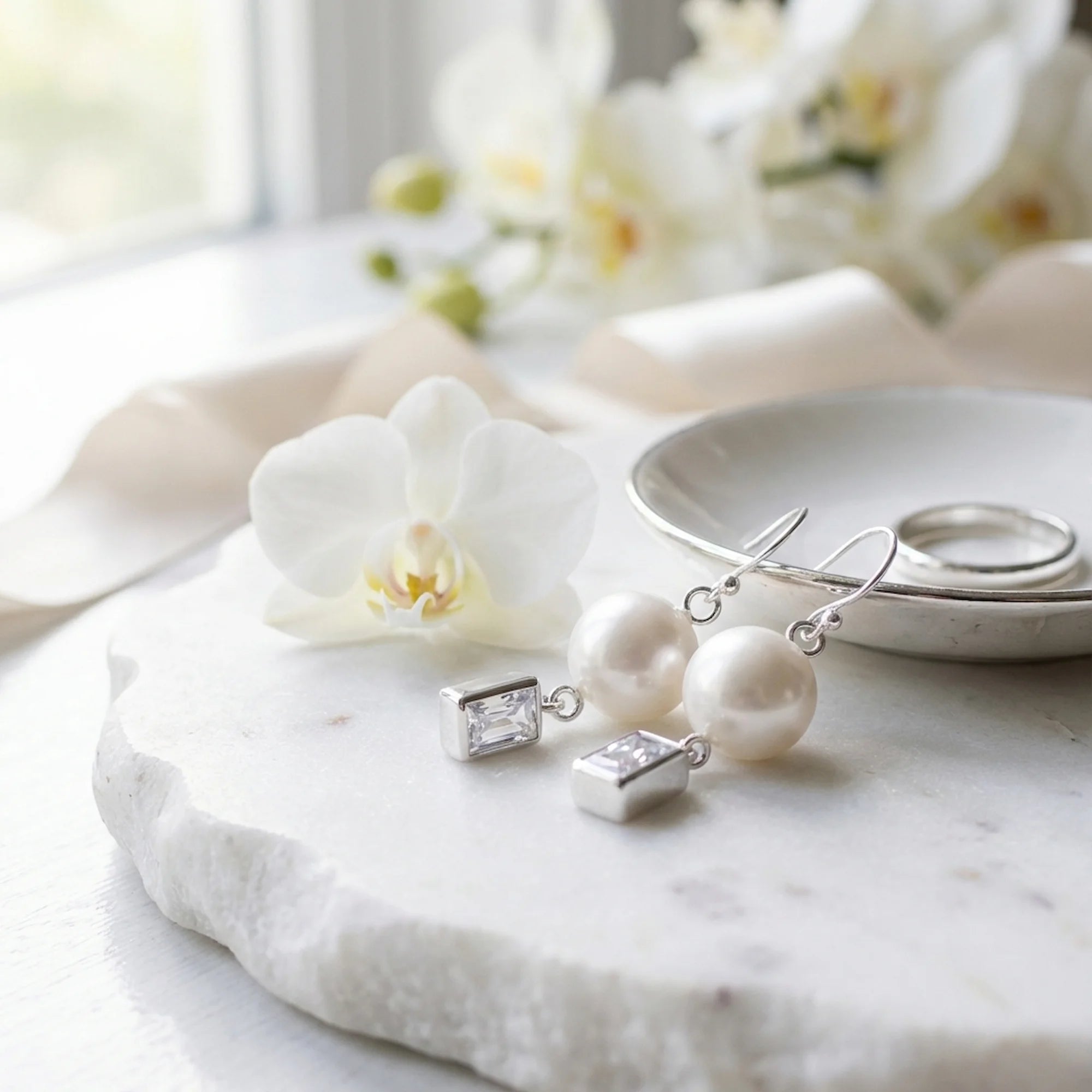 Silver pearl and rectangular cubic zirconia drop earrings beside a white orchid on marble
