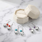 Three pairs of silver puffed heart bead earrings beside a cream velvet box on marble — red, blue and purple bead drops