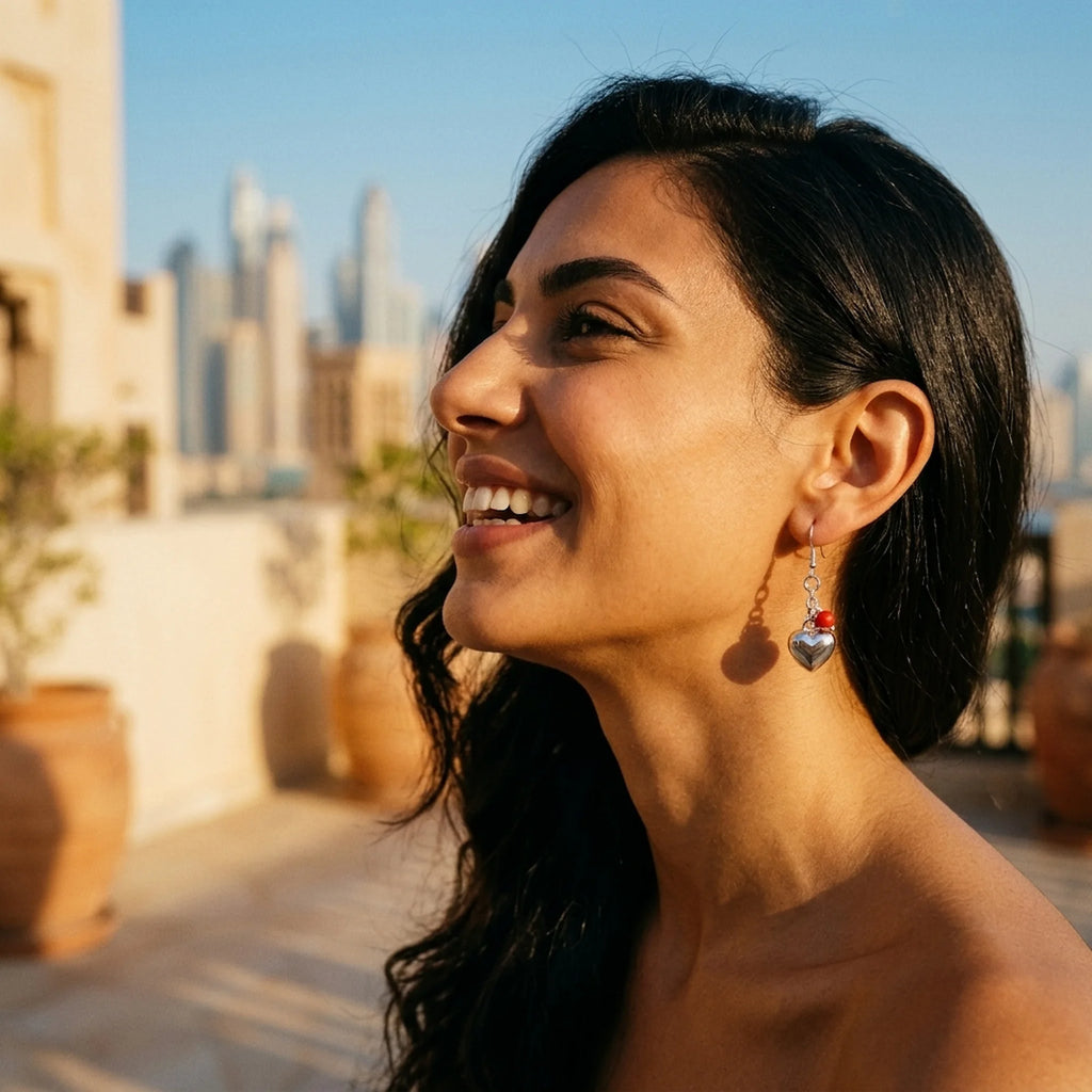 Silver puffed heart dangle earring with a red bead worn on the ear, Dubai skyline behind — 925 sterling silver drop