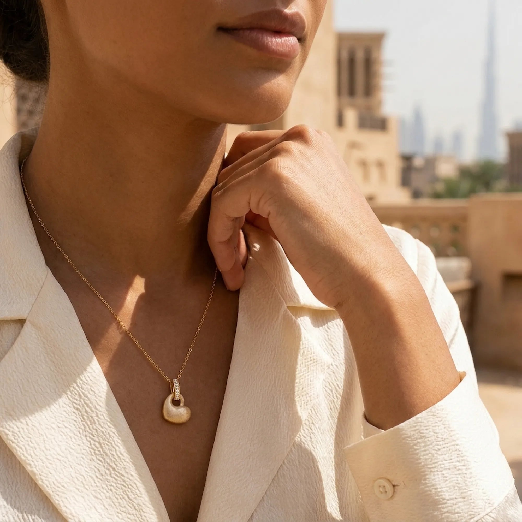 Matte gold puffed heart pendant necklace on olive skin model with Burj Khalifa Dubai backdrop — cream blazer