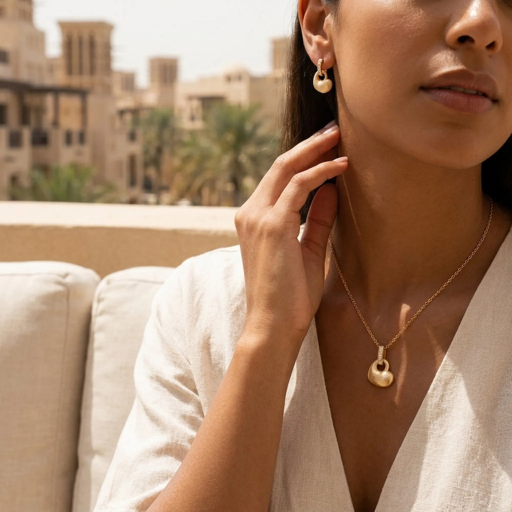 Satin gold puffed heart drop earrings and pendant set worn with white linen V-neck at Dubai outdoor terrace