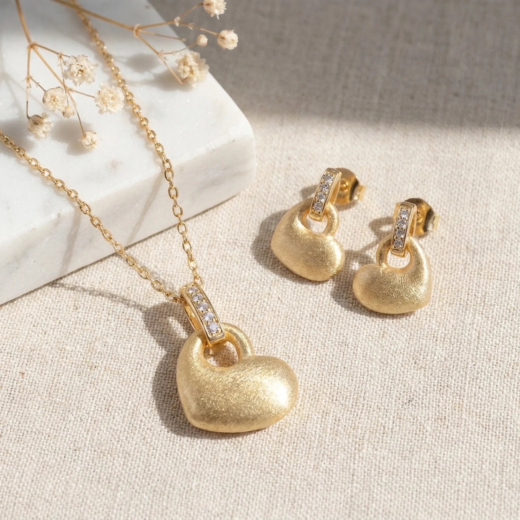 Brushed gold puffed heart pendant necklace and drop earring set on natural linen with dried flowers — gift display