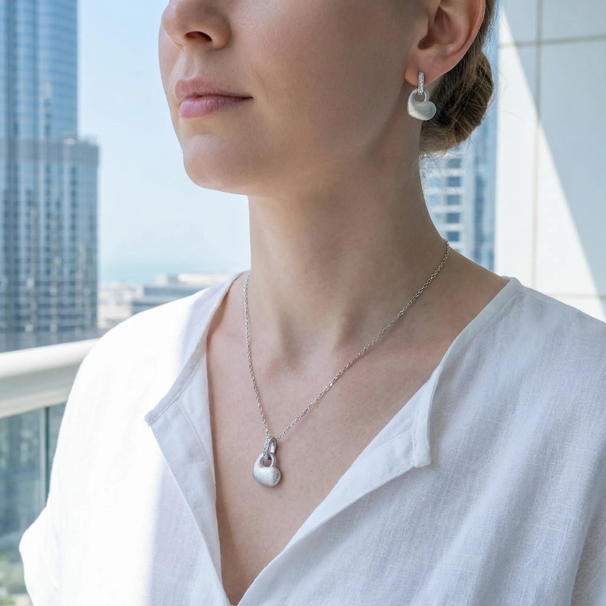 Brushed silver puffed heart earring and pendant set worn with white top against UAE modern architecture