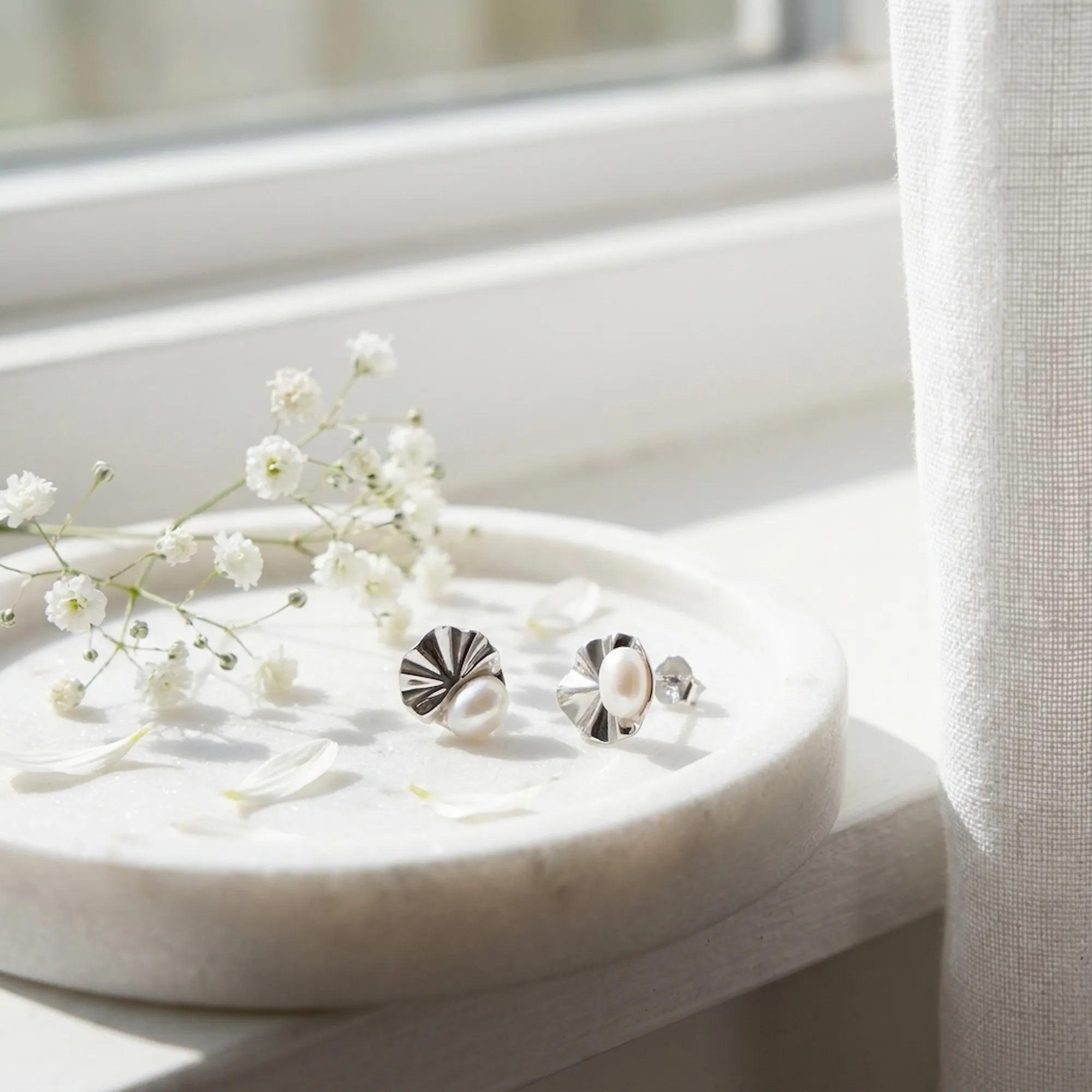 Shell pearl rhodium silver stud earrings on marble dish with white gypsophila flowers in natural light