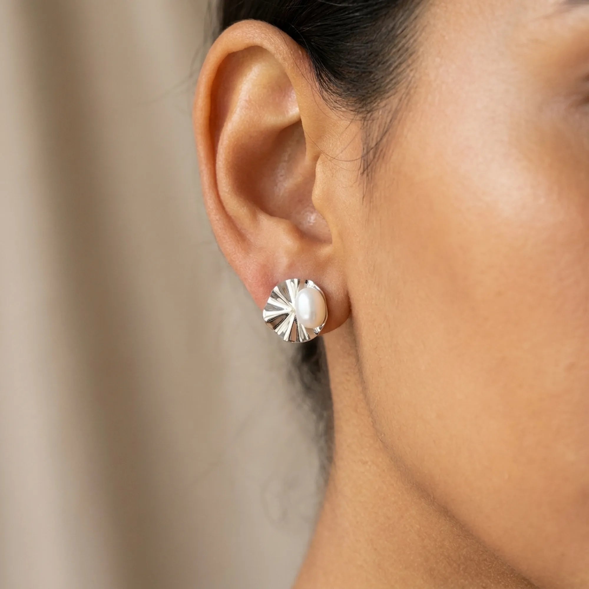 Close-up of shell pearl stud earring on ear against neutral draped fabric, rhodium fan shell detail
