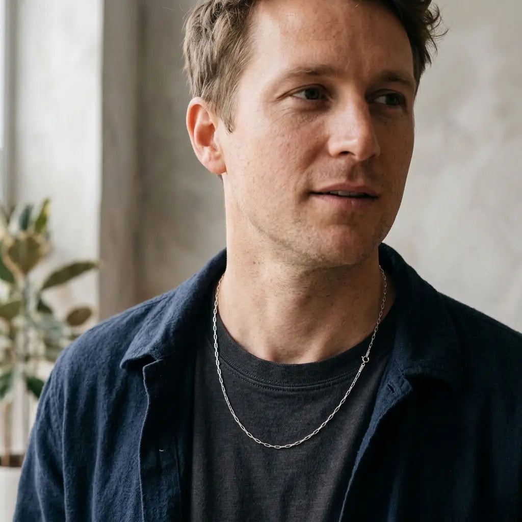 Man wearing a fine silver paperclip-link necklace over a dark T-shirt and navy overshirt.