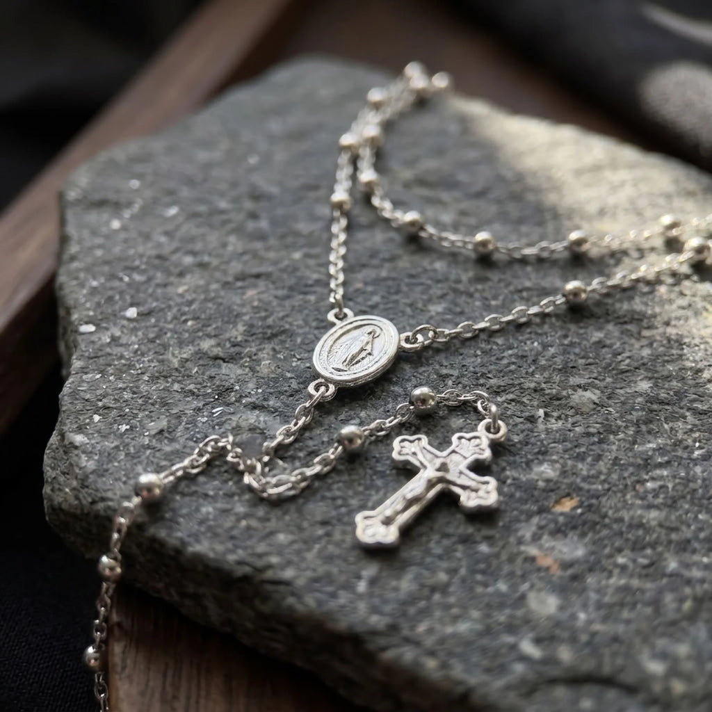 Close-up of Italian silver rosary-inspired necklace with Marian medal, polished beaded chain and detailed crucifix on dark stone.